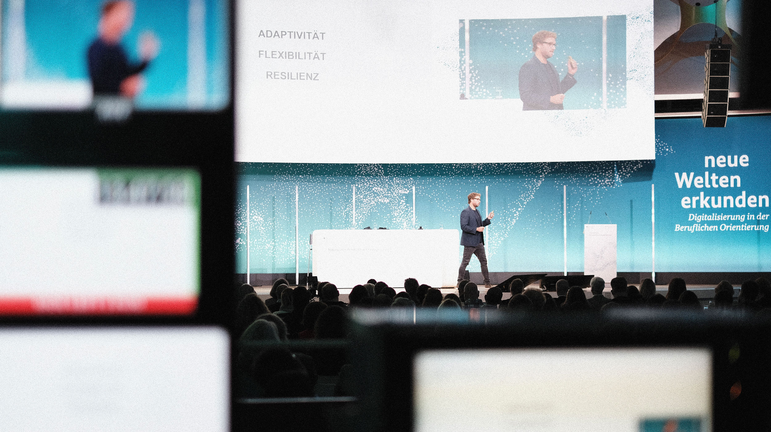 A speaker presents on stage at a conference, with a large screen displaying keywords: Adaptivität, Flexibilität, Resilienz.